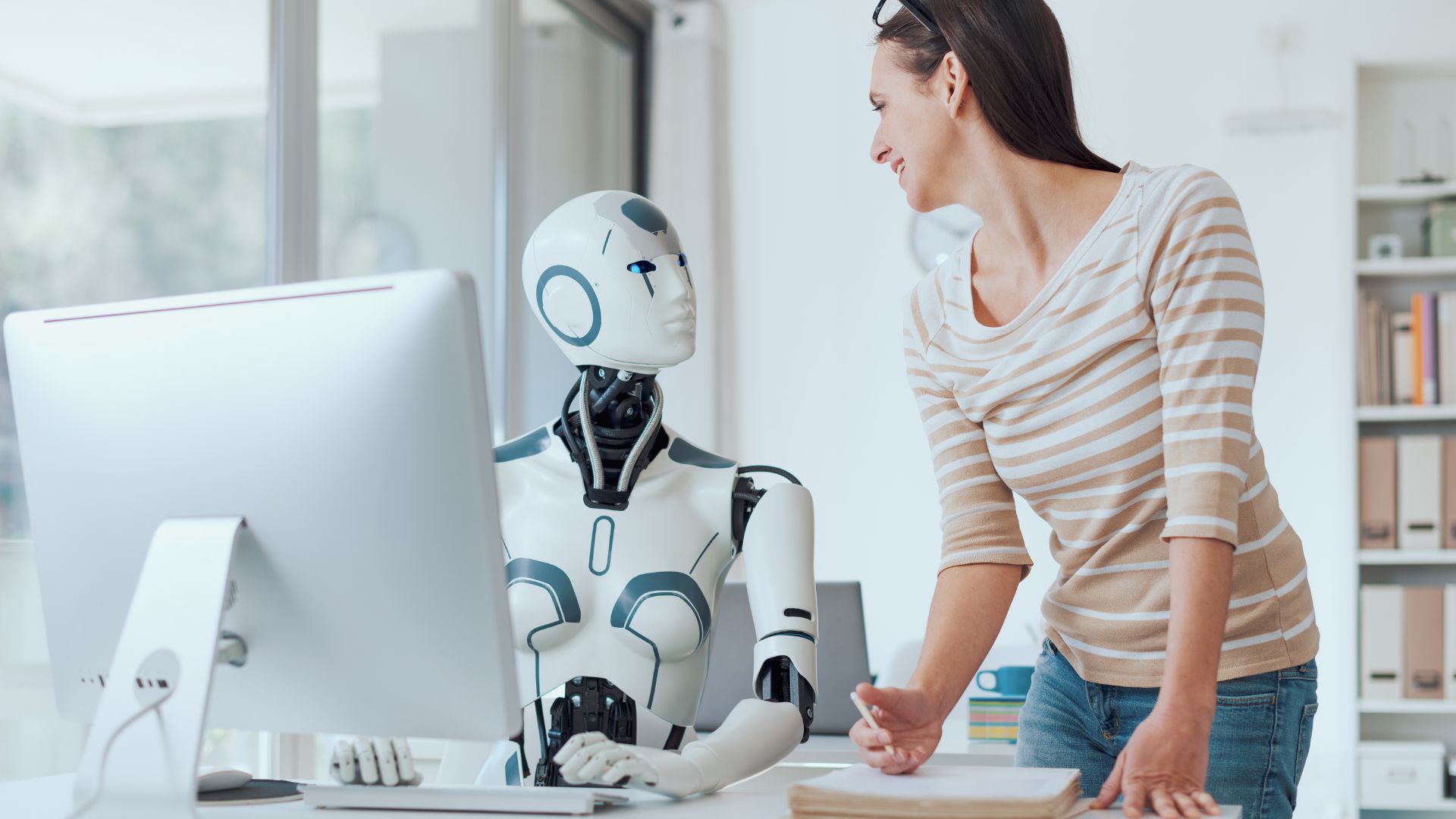AI-robot és ember közötti interakció, ahol a gép asszisztensként támogatja a felhasználót – ember-gép együttműködés a SailingHangar AI-leadership cikkéhez. AI robot and human collaborating at a computer – the machine assists the user in a co-pilot role, for SailingHangar’s AI leadership article.